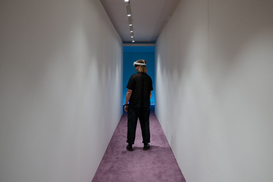 Photo from the Fields of Vision exhibition, showing a person wearing a VR headset walking through a hallway with purple carpet