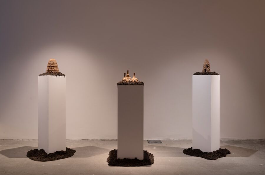 Photo from the Fields of Vision exhibition, showing an installation of three white plinths each with a clay sculptures on top of a pile of soil