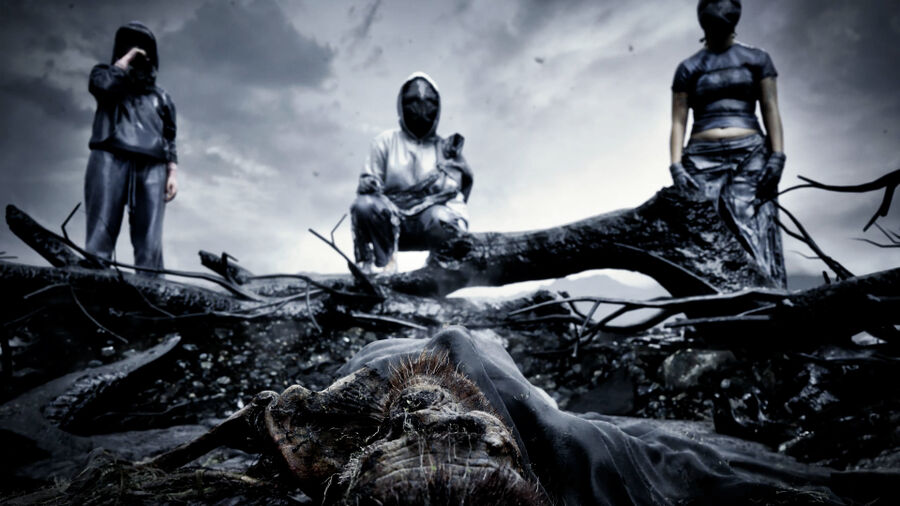 Still from the film The Bastard Fields, showing a bleak, post-apocalyptic scene with masked figures looming over a decayed corpse beneath a stormy sky.